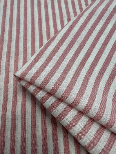 Red and white striped fabric is shown, with one piece neatly folded over itself, displaying a pattern of evenly spaced vertical stripes. The textured material suggests it's a woven blend of Linen Cotton - Pink Stripe - 145cm from Super Cheap Fabrics. The image focuses on the close-up details of the breathable fabric's design and texture.