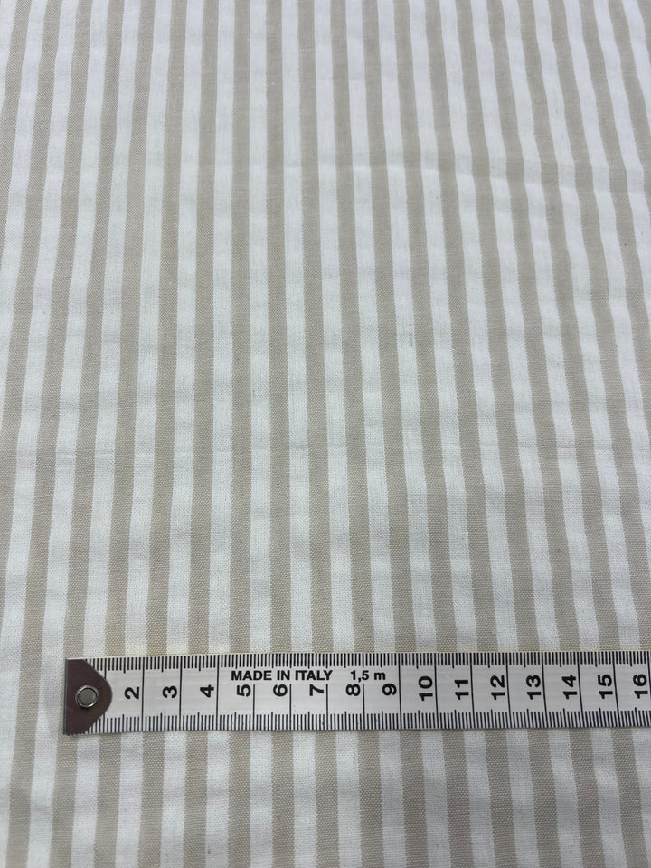 A piece of beige and white striped Linen Cotton - Arctic Wolf Stripe - 145cm from Super Cheap Fabrics, made from natural fibers like Linen Cotton, is measured with a tape measure marked in centimeters and millimeters. The measure displays "MADE IN ITALY," capturing the vertical, evenly spaced stripes.