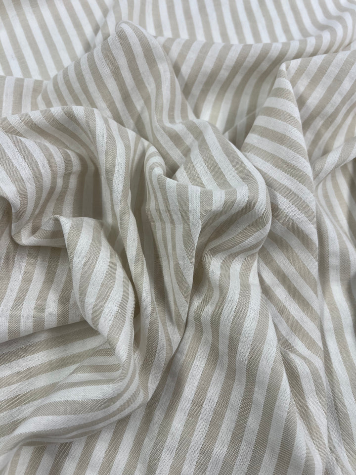 A close-up of a piece of fabric with a beige and white striped pattern. The Super Cheap Fabrics Linen Cotton - Arctic Wolf Stripe - 145cm appears soft and slightly crumpled, creating folds and shadows that add texture to the image. This lightweight fabric showcases the beauty of natural fibers in its intricate design.