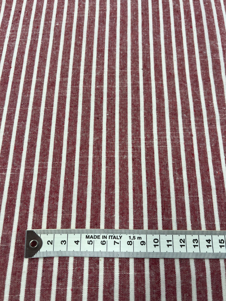 A piece of red and white vertically striped fabric made from Linen Cotton - Earth Red Stripe - 145cm by Super Cheap Fabrics is shown, with a white measuring tape placed horizontally at the bottom. The measuring tape indicates a length of 14 centimeters and has the text "MADE IN ITALY" on it.