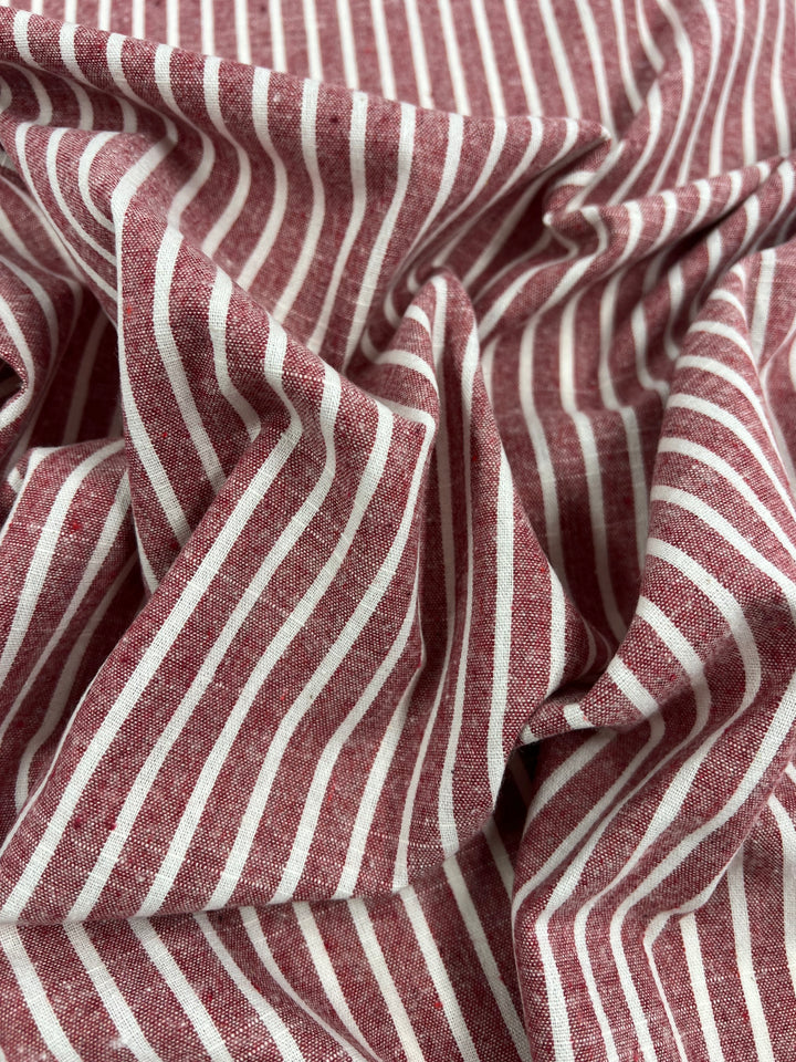 Close-up of crumpled fabric featuring a pattern of white and burgundy vertical stripes. The Super Cheap Fabrics Linen Cotton - Earth Red Stripe - 145cm appears textured, with a slightly wrinkled look, highlighting the breathable nature of the natural fibers.
