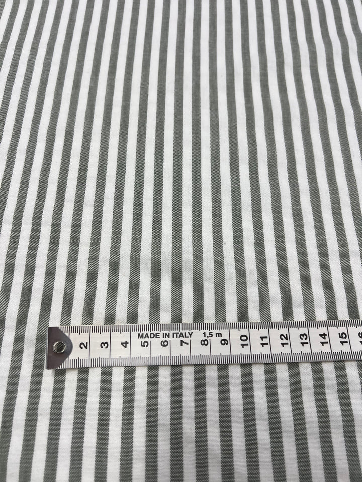 A close-up image of a Linen Cotton - Alfalfa Stripe - 145cm fabric from Super Cheap Fabrics with vertical gray and white stripes. A metal measuring tape, displaying both centimeter and inch markings, is placed horizontally across the natural fibers near the bottom of the image. The tape reads "Made in Italy.