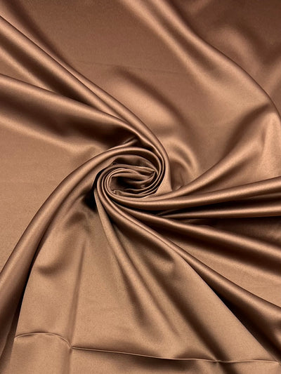 A smooth, shiny Satin Deluxe - Mocha Mousse - 150cm from Super Cheap Fabrics is elegantly twisted into a swirl pattern. The rich texture and reflective surface of this luxury fabric create a luxurious and soft appearance, highlighting the fabric's fluidity and delicate folds.