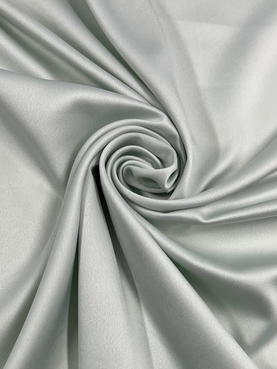 A close-up of Super Cheap Fabrics' Satin Deluxe - Frost - 150cm arranged in a smooth, swirling pattern. The folds create a spiral design at the center, showcasing the material's soft and silky texture with a shiny finish.