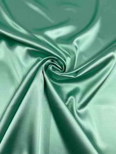 A neatly arranged piece of shiny, emerald green satin fabric with smooth, soft folds gathered at the center, creating a luxurious swirl pattern. The fabric reflects light, highlighting its smooth texture and glossy finish, reminiscent of opulent nightgowns. This is Satin Deluxe - Honeydew - 150cm by Super Cheap Fabrics.