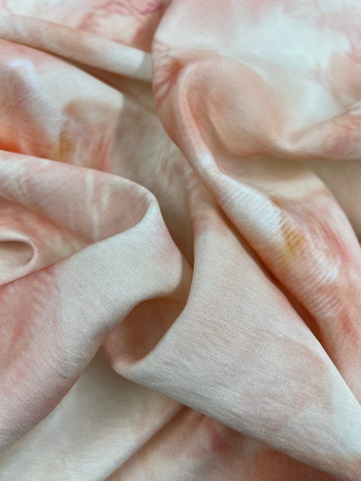 A close-up image of soft, crumpled children's clothing featuring a light pastel color palette. The main hues include gentle shades of pink, peach, and creamy white, creating a delicate and flowing appearance. The smooth texture of the lightweight fabric is accentuated by gentle folds and creases. This beautiful fabric is Designer Cotton - TieFly - 145cm from Super Cheap Fabrics.