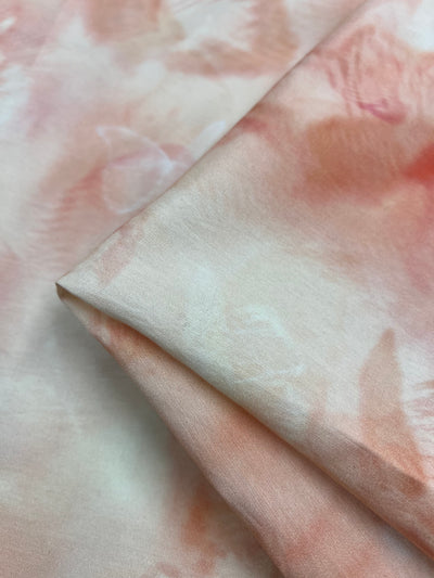 A close-up of soft, silky fabric with a gentle tie-dye pattern in pastel pink and peach tones. Ideal for children's clothing, the light weight fabric is slightly folded, showcasing its smooth texture and subtle color variations. This is Designer Cotton - TieFly - 145cm by Super Cheap Fabrics.