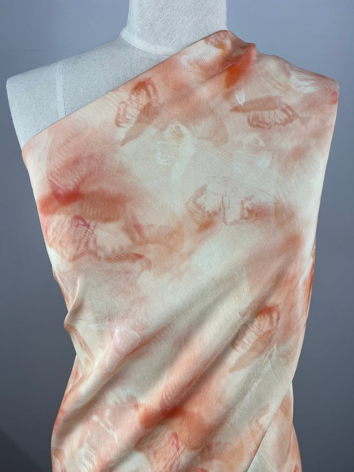 A section of Designer Cotton - TieFly - 145cm by Super Cheap Fabrics draped over a white mannequin, featuring a soft, abstract tie-dye pattern with shades of pale peach, orange, and cream. Perfect for children's clothing, the background is a plain gray, allowing the warm colors of the fabric to stand out.