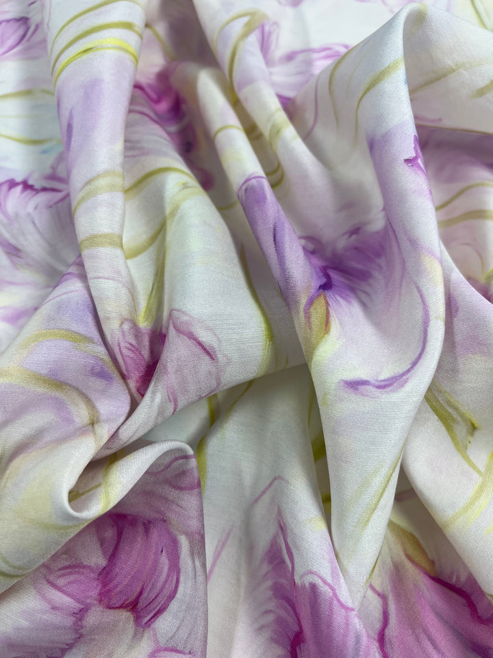 A close-up of soft, draped fabric featuring an abstract floral pattern in pastel hues of purple, pink, and light green against a white background. This lightweight fabric has a smooth texture with delicate flowing lines, making it ideal for children's clothing and other multi uses. The product is Designer Cotton - Peony Dreams - 145cm by Super Cheap Fabrics.