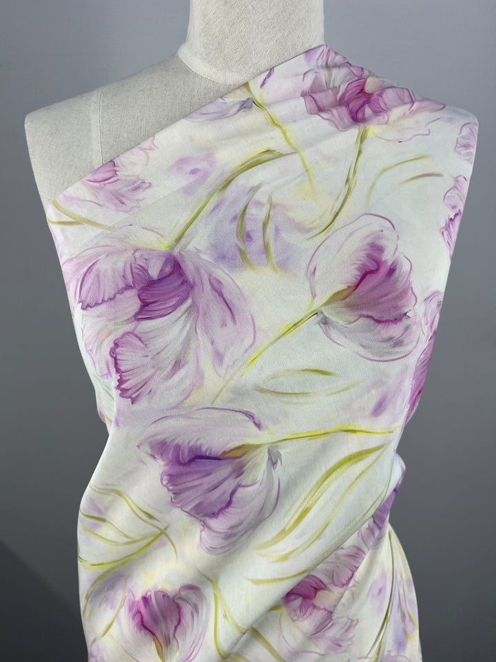 A light weight Designer Cotton - Peony Dreams - 145cm draped over a mannequin, featuring a floral pattern with light purple flowers and green stems on a light background. The fabric appears to be made of soft, Super Cheap Fabrics' product material, giving it an elegant and delicate look—ideal for children's clothing or other applications.