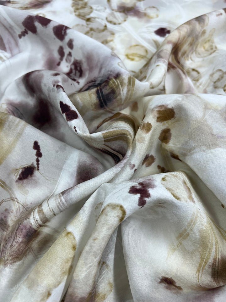A close-up of a piece of Super Cheap Fabrics Designer Cotton - Bubbles - 145cm with an abstract, watercolor-like pattern. The design, ideal for children's clothing, features soft, flowing folds and a palette of muted colors, including shades of brown, beige, and touches of purple. The texture appears smooth and slightly reflective.