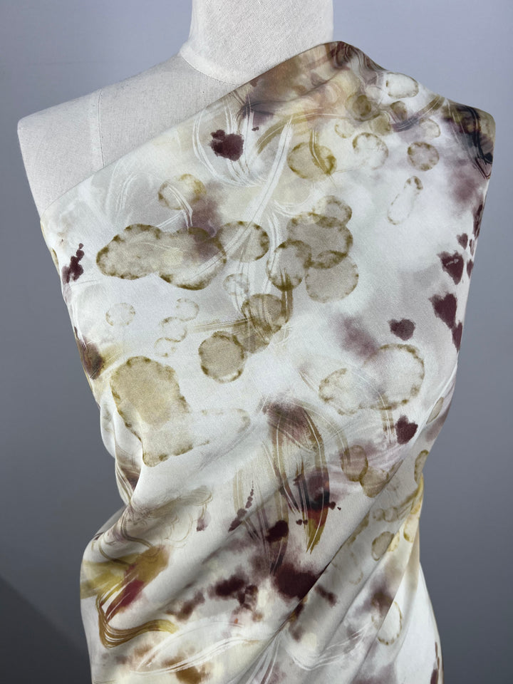 A mannequin is draped with Designer Cotton - Bubbles - 145cm by Super Cheap Fabrics, a lightweight fabric featuring abstract patterns in shades of brown, beige, and light purple. The multi-color fabric design includes blotchy, cloud-like formations with swirls and splatters, giving it an artistic, watercolor-like appearance.
