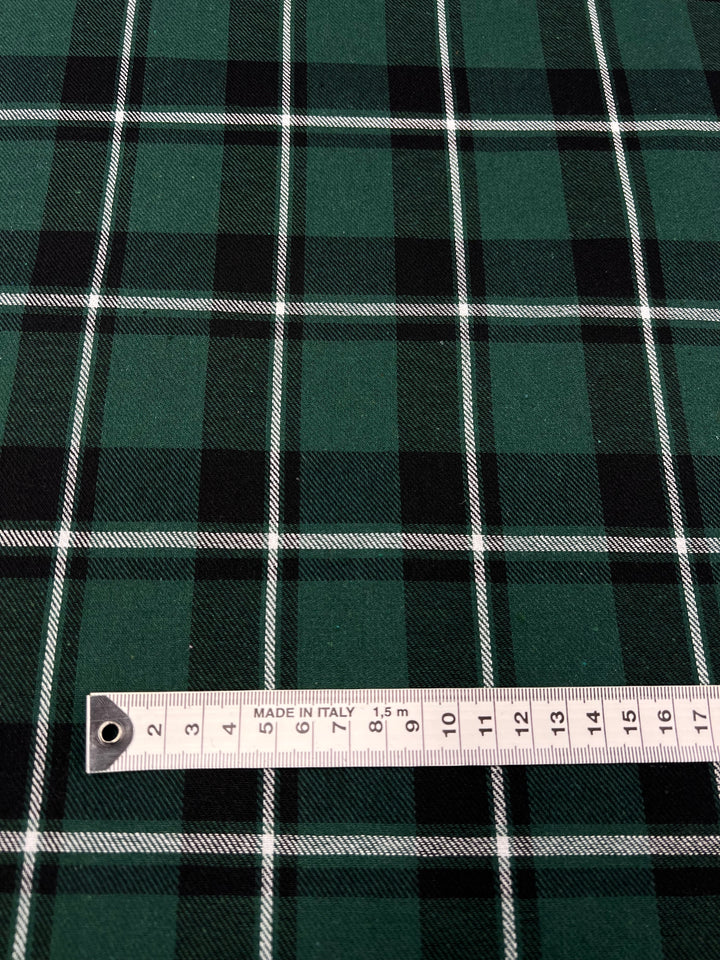 A close-up view of a green and black tartan twill suiting fabric with white lines forming a plaid pattern. A measuring tape marked "Made in Italy" lies horizontally across the versatile garment, showing measurements ranging from 2 to 15 centimeters. The fabric is the Suiting - Emerald Tattersal - 150cm by Super Cheap Fabrics.