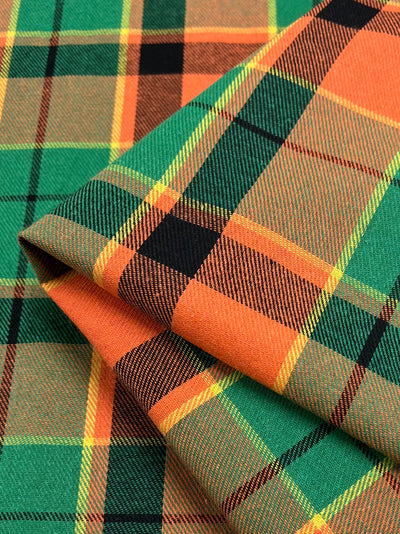 A close-up of folded suiting fabric displaying a colorful tartan pattern. The design features intersecting stripes in green, orange, yellow, and black, creating a vibrant and traditional checkered lattice. The lightweight fabric appears textured and woven. This is the Suiting - Citrus Tartan - 150cm by Super Cheap Fabrics.