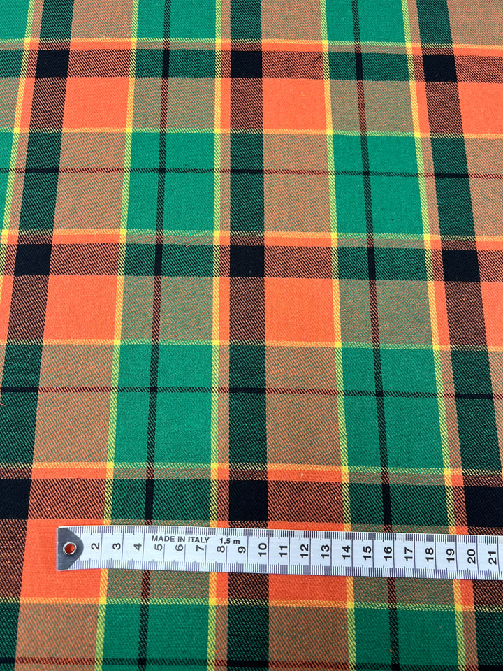 Close-up of a colorful plaid suiting fabric featuring orange, green, yellow, and black stripes in a crisscross pattern. A metal ruler placed on the lightweight Suiting - Citrus Tartan - 150cm by Super Cheap Fabrics indicates it is made in Italy and measures up to 15 centimeters.