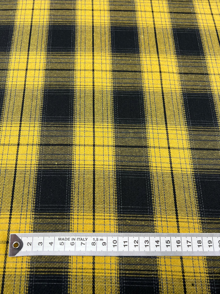 A close-up view of a versatile fabric with a yellow and black plaid pattern. A measuring tape is laid out horizontally at the bottom of the image, showing markings from 0 to 20 centimeters. The wrinkle-resistant fabric, Suiting - Clueless - 150cm by Super Cheap Fabrics, has a "Made in Italy" label.