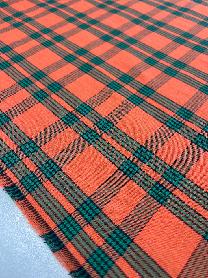 Close-up of a versatile fabric featuring a plaid pattern with vibrant orange, green, and black stripes. The threads are tightly woven, creating a checkerboard effect reminiscent of Suiting - Tartan Mandarin - 150cm by Super Cheap Fabrics. The fabric appears to be spread out flat, with the edge of the material visible on the left side.