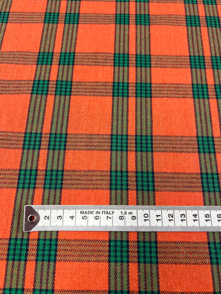 A close-up of orange, green, and brown plaid fabric with a metal ruler placed horizontally across it. The ruler, marked with centimeters, shows a length of 16 cm and includes the text "MADE IN ITALY." This versatile fabric from Super Cheap Fabrics is ideal for formal suits or twill suiting applications and is called Suiting - Tartan Mandarin - 150cm.