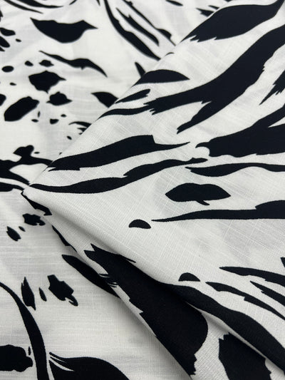 A close-up photo of a fabric with a white background and abstract black patterns. The Super Cheap Fabrics Designer Bamboo Rayon - Liquid Zebra - 147cm appears folded, showcasing its smooth texture and bold, irregular designs reminiscent of brushstrokes or animal print. This versatile choice features vibrant prints perfect for any creative project.