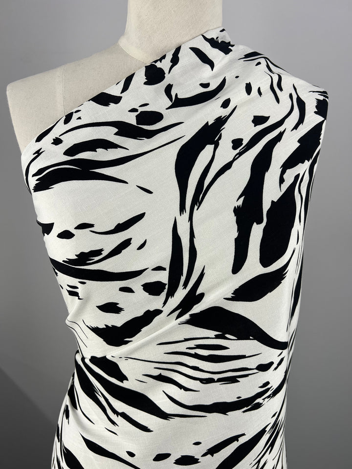 A mannequin draped in a one-shoulder, black and white abstract print Designer Bamboo Rayon - Liquid Zebra - 147cm by Super Cheap Fabrics. The bold pattern features sweeping black strokes and shapes on a white background, making it a versatile choice. The photo focuses on the upper torso and part of the shoulder.