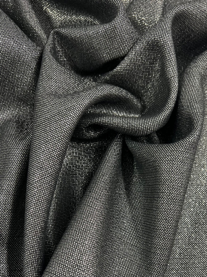 Close-up of a dark, glossy, textured fabric with a metallic sheen. The fabric appears to be draped and gathered, creating folds and creases that reflect light. Metallic silver threads woven into this Wool Lamé - Metallic - 150cm by Super Cheap Fabrics add to its shimmery appearance.