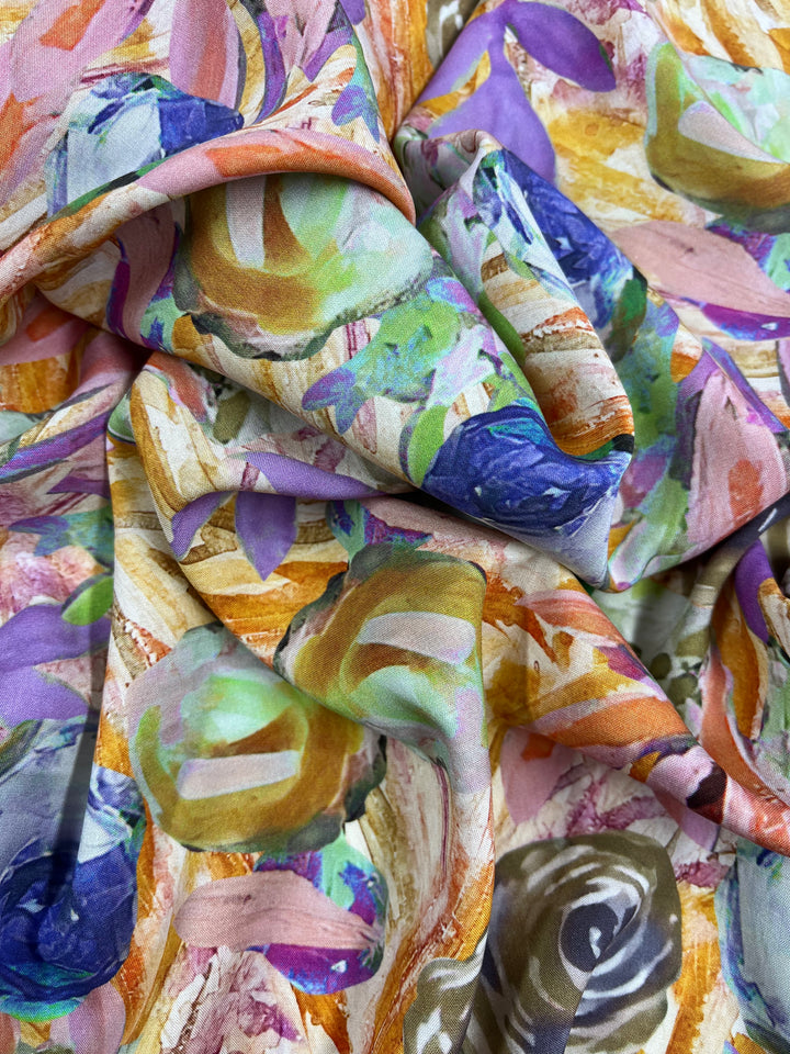 A close-up of vibrant, multi-colored fabric draped in folds. The Super Cheap Fabrics Designer Rayon - Nursery Dreams - 145cm features an abstract pattern with swirls and splashes of purple, green, orange, and blue hues, creating a dynamic and textured appearance. This versatile fabric appears soft and lightweight.