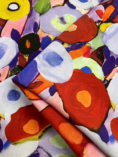 A close-up of a vibrant, colorful fabric featuring an abstract floral pattern. This versatile fabric, made from Super Cheap Fabrics' Designer Rayon - Poppy Fields - 145cm, showcases bold colors like red, purple, yellow, green, and blue in an artistic painterly style. The texture and folds of the vibrant print are visible in the image.