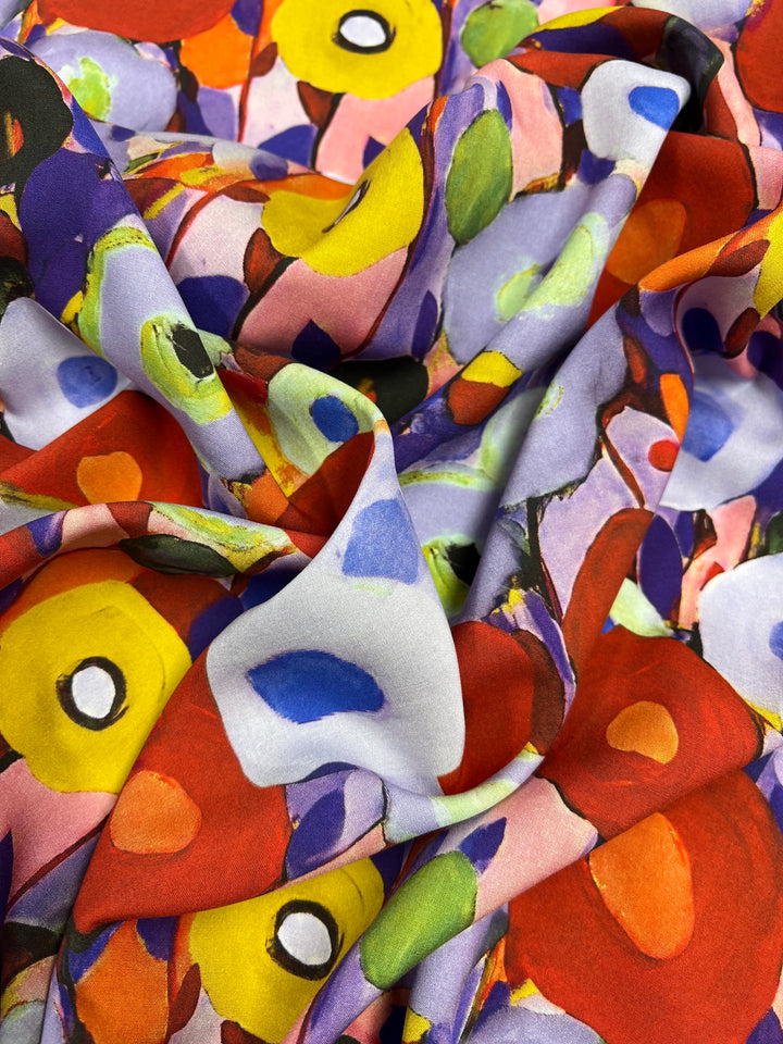 A close-up image of Super Cheap Fabrics Designer Rayon - Poppy Fields - 145cm with a colorful, abstract floral pattern. The print features large, vibrant flowers in shades of red, yellow, green, purple, and blue, with bold black and white accents. The versatile fabric appears to be draped or crumpled.