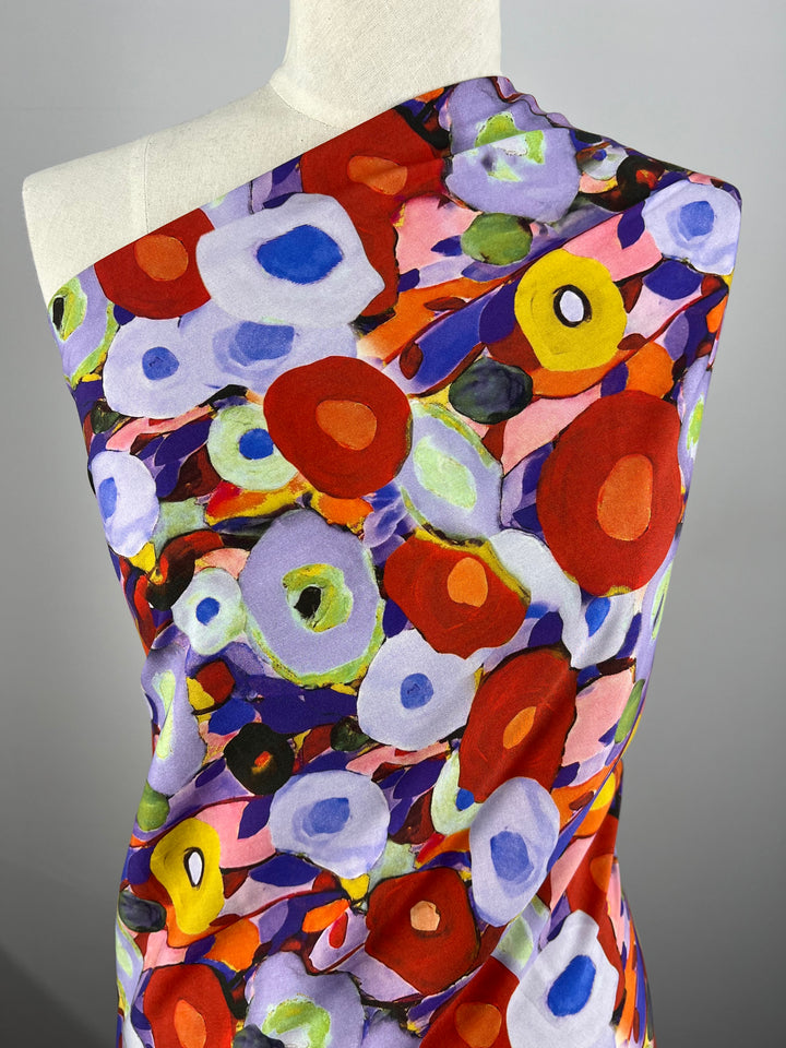 A mannequin is draped in a vividly colored Super Cheap Fabrics Designer Rayon - Poppy Fields - 145cm featuring an abstract floral pattern. The design includes red, orange, yellow, purple, blue, and green circular flower shapes on a multi-colored background, creating a lively and artistic visual effect.
