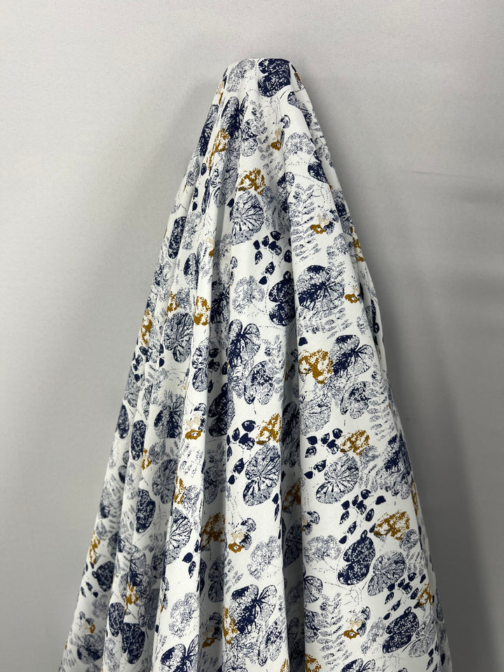 A piece of white, light weight fabric draped against a wall, featuring an intricate pattern of blue and gold abstract prints, including floral and leaf motifs. Crafted from 100% cotton, the **Cotton Sateen - Lily Pad - 145cm** by **Super Cheap Fabrics** has a slightly glossy finish and the detailed designs are scattered throughout, creating a textured effect.