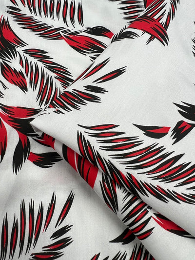 A close-up of a light weight, 100% cotton fabric with a dynamic pattern consisting of red, black, and white colors. The design has a brushstroke-like effect that creates an abstract, feathered appearance. Perfect for dresses, the fabric is slightly folded, adding texture and depth to the image. This is Cotton Sateen - Palm Stroke - 140cm from Super Cheap Fabrics.