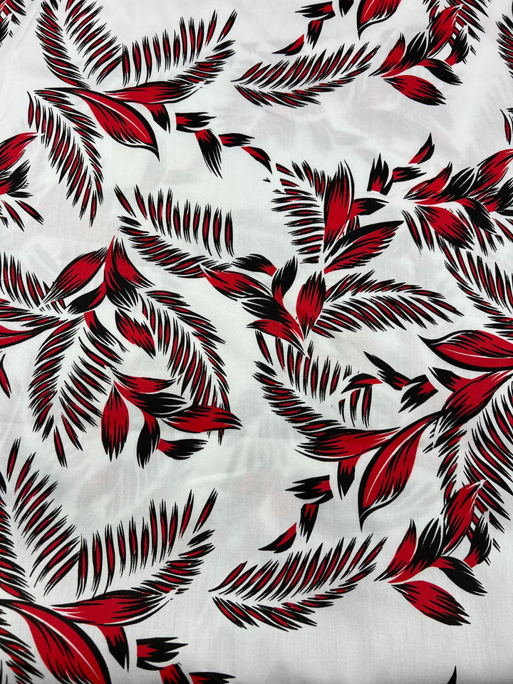 A light weight fabric with a white background featuring a repeating pattern of red and black abstract palm leaf designs. Made from 100% cotton, the leaves are various sizes and oriented in different directions, creating a dynamic and eye-catching effect perfect for dresses. This is Super Cheap Fabrics' Cotton Sateen - Palm Stroke - 140cm.