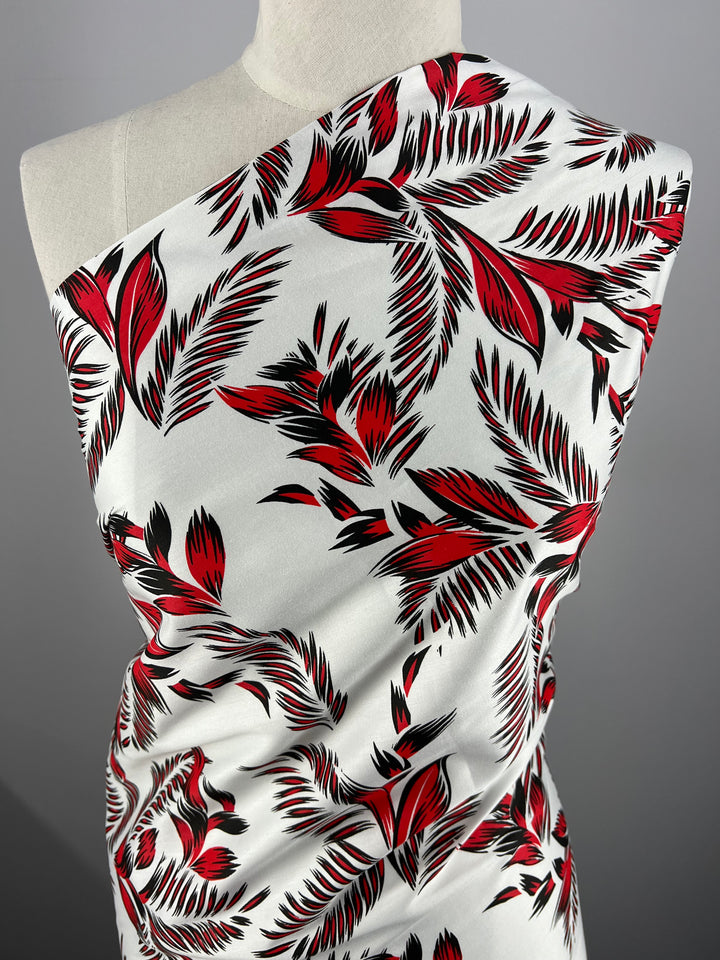 A white fabric drapes a mannequin, featuring a vibrant red and black tropical leaf pattern. The 100% cotton material showcases various palm leaves and abstract shapes, creating a bold and elegant appearance. The light weight fabric's flowing texture is highlighted under soft lighting, ideal for dresses. This is the Cotton Sateen - Palm Stroke - 140cm by Super Cheap Fabrics.