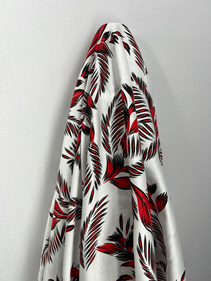 A lightweight, 100% cotton fabric with a bold, red and black tropical leaf pattern draped against a plain white background. The design features various leaf shapes, creating a dynamic and vibrant visual effect perfect for dresses. This is the Cotton Sateen - Palm Stroke - 140cm from Super Cheap Fabrics.