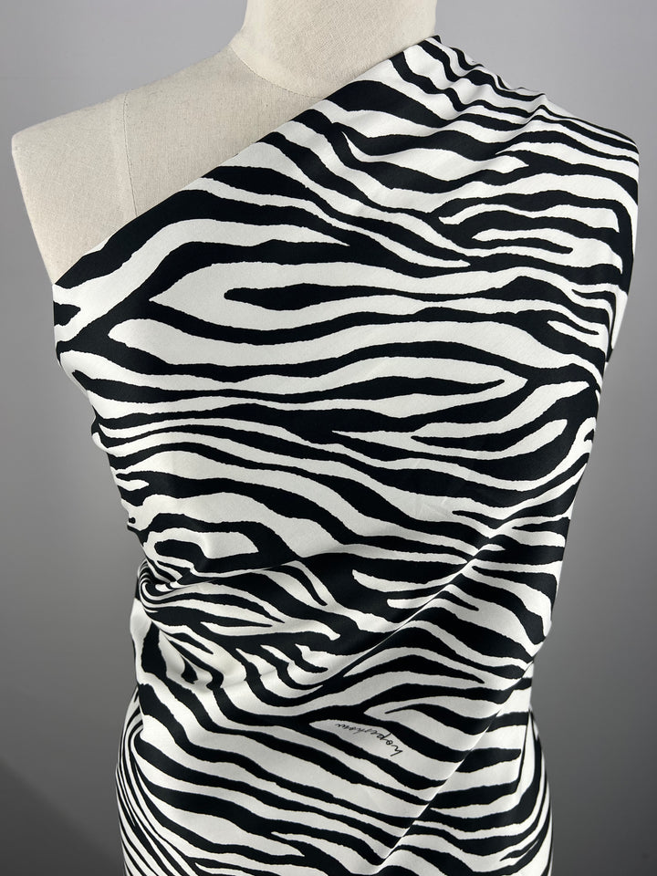 A mannequin is draped with light weight fabric featuring a striking black and white zebra print pattern. Made from 100% cotton, the material covers one shoulder and flows diagonally across the torso. The plain gray background puts full focus on this bold design, perfect for household décor projects. This is Cotton Sateen - Zebray - 150cm by Super Cheap Fabrics.