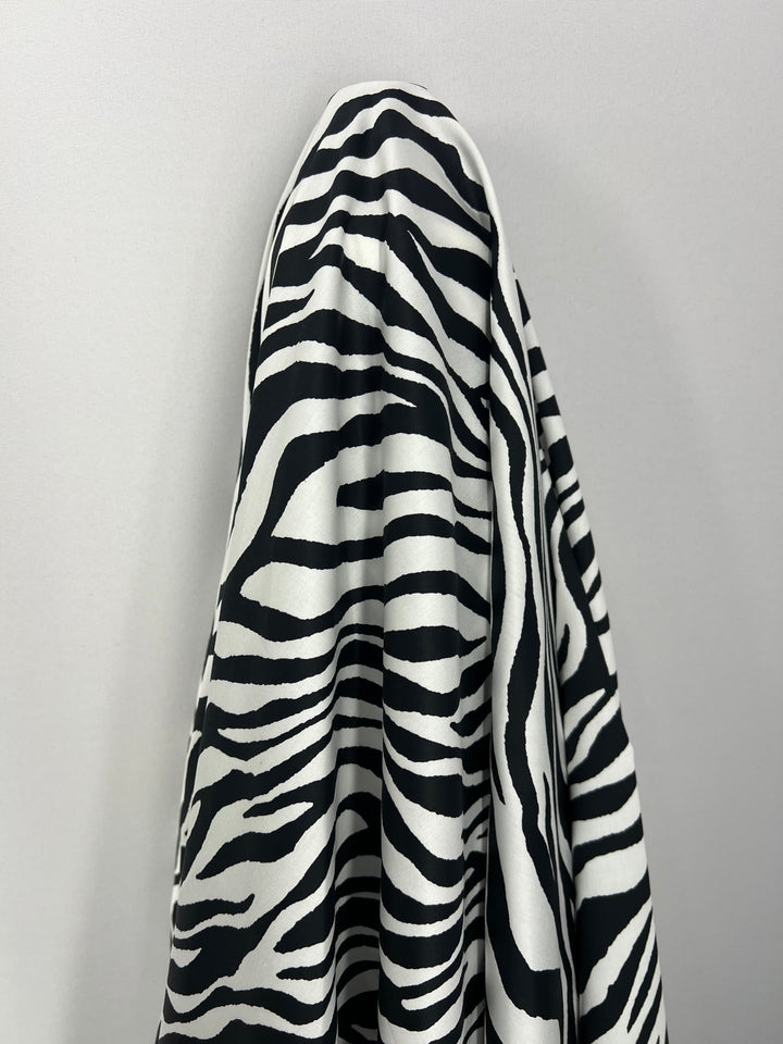 A piece of light weight fabric with a black and white zebra stripe pattern is draped over a hook or object against a plain white background. The 100% cotton fabric, Cotton Sateen - Zebray - 150cm by Super Cheap Fabrics, appears to be silky and is hanging in folds, perfect for household décor.
