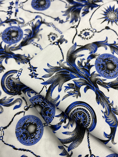 A close-up image of light weight fabric with intricate blue and black floral and geometric patterns on a white background. The 100% cotton design features detailed circular motifs connected by delicate chains, giving the fabric an elegant and ornate appearance, perfect for dresses. This is Cotton Sateen - Compass - 145cm by Super Cheap Fabrics.