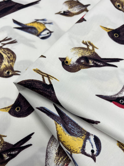 A close-up of a folded piece of bird print fabric featuring a colorful design. The birds, in shades of brown, yellow, and blue, are scattered across a white background, depicted in various poses. This Super Cheap Fabrics Cotton Sateen - Sanctuary - 150cm showcases their intricate details beautifully.