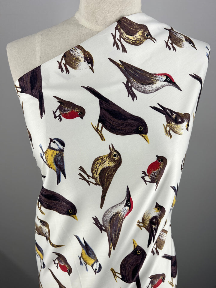 A close-up of bird print fabric draped over a mannequin, showcasing various birds in different poses and colors, including red, yellow, brown, and white, set against a light background. This is the Cotton Sateen - Sanctuary - 150cm from Super Cheap Fabrics.