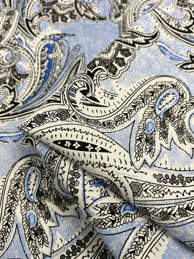 A close-up image of Cotton Sateen - Paisley Baroque - 145cm from Super Cheap Fabrics with an intricate paisley pattern. The design features curved, teardrop-shaped figures in black and white with blue accents on a light blue background. The lightweight fabric appears folded, emphasizing the texture and detail of the print, perfect for household décor.