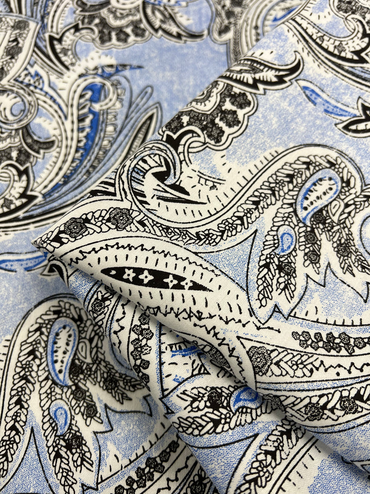 A close-up image of Cotton Sateen - Paisley Baroque - 145cm from Super Cheap Fabrics with an intricate paisley pattern. The design features curved, teardrop-shaped figures in black and white with blue accents on a light blue background. The lightweight fabric appears folded, emphasizing the texture and detail of the print, perfect for household décor.