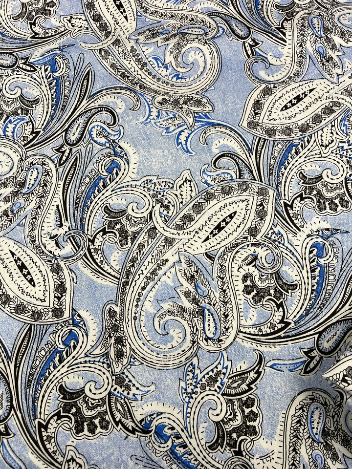 A detailed paisley pattern featuring swirling, intricate designs in blue, black, and white shades. The background consists of a light blue color, giving the Super Cheap Fabrics Cotton Sateen - Paisley Baroque - 145cm an elegant and classic appearance perfect for household décor.
