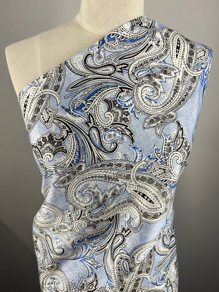 A mannequin draped in a single-shoulder garment with a paisley pattern in shades of blue, black, and white. The intricate design features swirling, leaf-like shapes and detailed line work on light-weight fabric made from 100% cotton. The fabric used is "Cotton Sateen - Paisley Baroque - 145cm" by Super Cheap Fabrics.