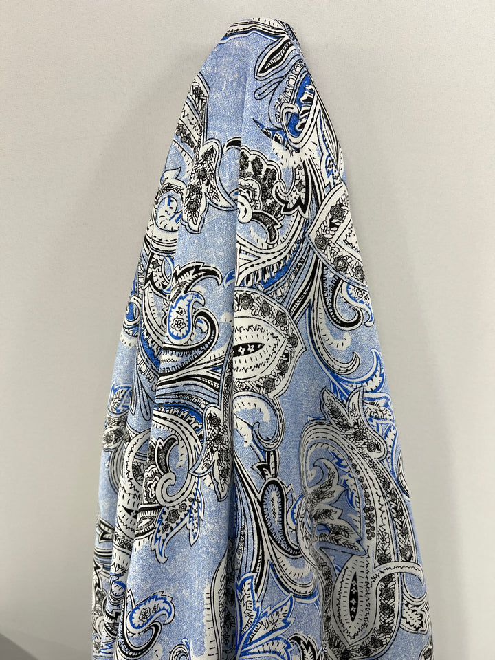 A **Super Cheap Fabrics Cotton Sateen - Paisley Baroque - 145cm** curtain with an intricate paisley pattern in shades of blue, black, and white is draped against a plain light gray wall, adding a touch of elegance to your household décor.