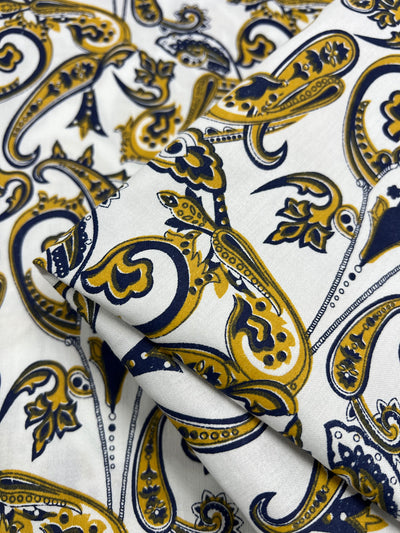 Close-up image of Super Cheap Fabrics' Cotton Sateen - Paisley Swirl - 140cm with a paisley pattern in yellow, navy blue, and black on a white background. The intricate design on this lightweight, 100% cotton material features large, ornamental teardrop shapes with detailed floral and abstract elements, creating a vibrant and sophisticated look.