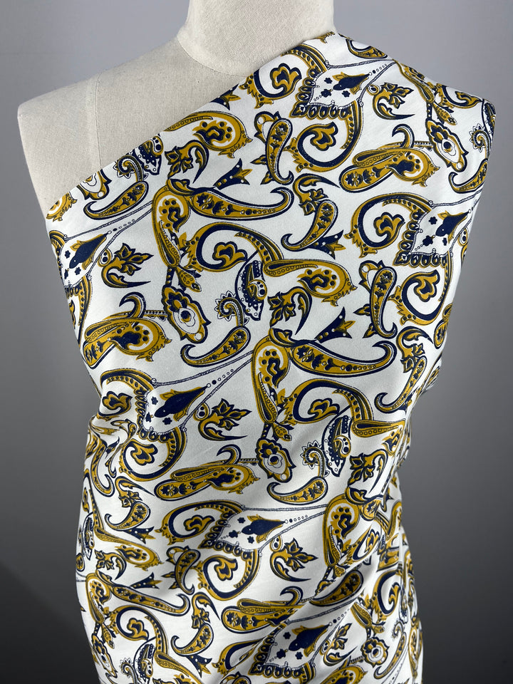 A mannequin is draped with Super Cheap Fabrics' Cotton Sateen - Paisley Swirl - 140cm featuring an intricate paisley pattern in yellow, blue, and black on a white background. The 100% cotton fabric covers one shoulder of the mannequin, showcasing the elaborate design. Ideal for household décor.