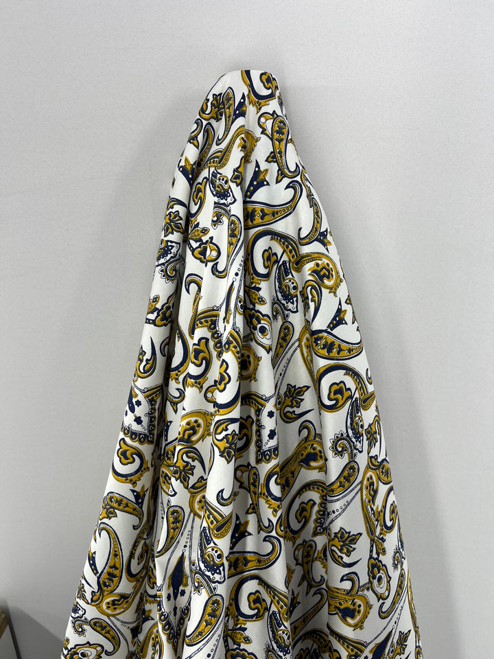 A piece of Cotton Sateen - Paisley Swirl - 140cm by Super Cheap Fabrics hangs against a light gray background. The lightweight fabric features a vibrant paisley pattern in shades of yellow, navy blue, and white, with intricate, flowing designs creating an elegant and colorful appearance—perfect for household décor.