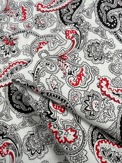 A close-up of a fabric featuring an ornate paisley pattern in black, white, and red. The intricate design includes various floral and curvilinear elements. Made from 100% cotton, the lightweight fabric is slightly crumpled, showcasing its texture and detail—perfect for household décor. This is the Cotton Sateen - Persian Pickles - 140cm from Super Cheap Fabrics.
