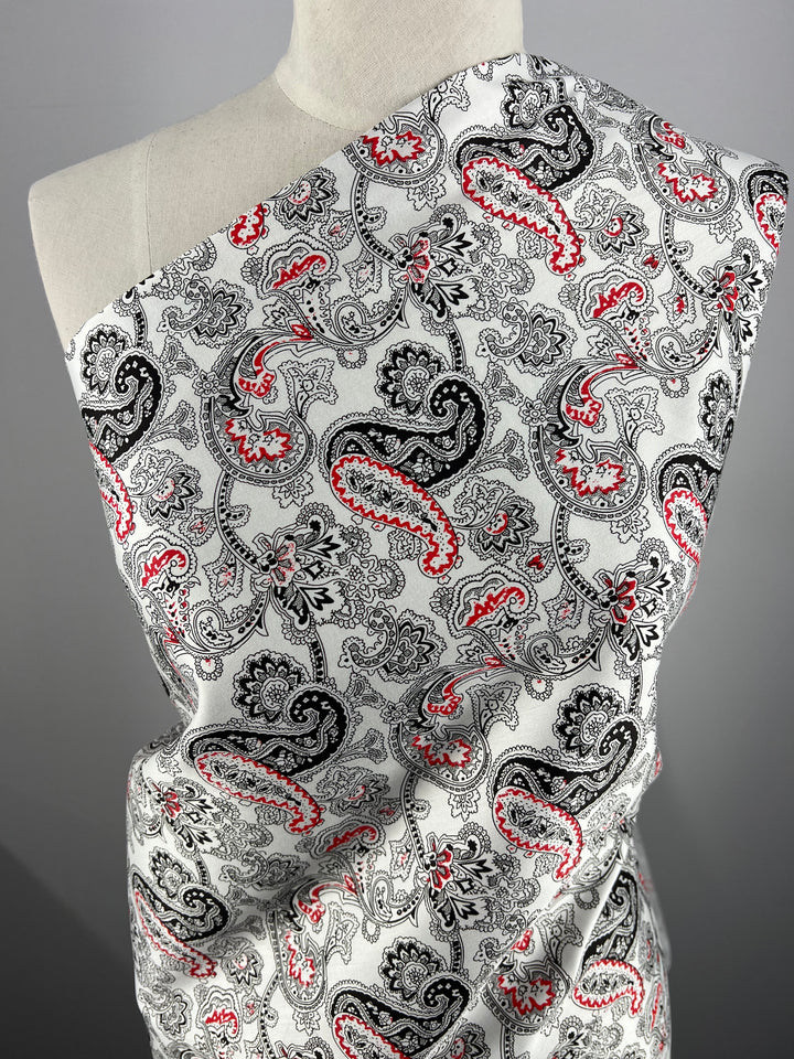 A mannequin draped with Super Cheap Fabrics' Cotton Sateen - Persian Pickles - 140cm featuring a detailed paisley pattern in black, white, and red colors. The intricate design includes swirling motifs and floral elements on a light background, perfect for household décor.