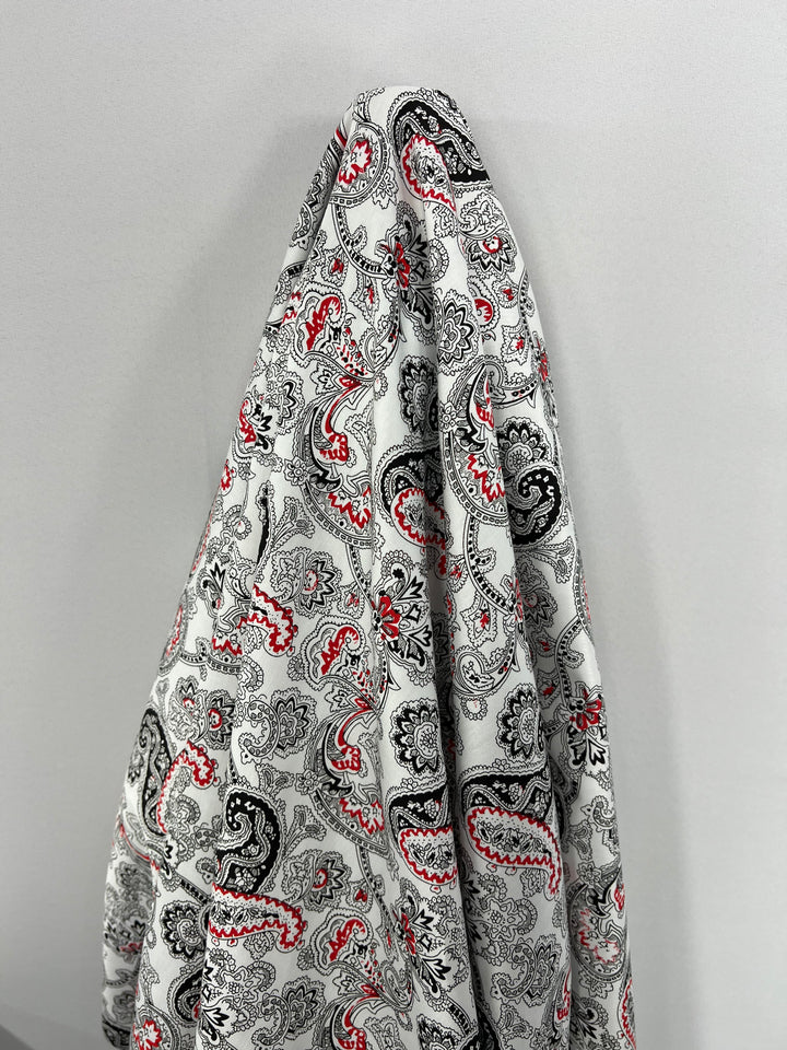 A piece of light-weight fabric with a paisley pattern hangs against a plain, light-colored wall. This *Super Cheap Fabrics* **Cotton Sateen - Persian Pickles - 140cm** features intricate designs in black, red, and shades of gray on a white background, making it perfect for elegant household décor.